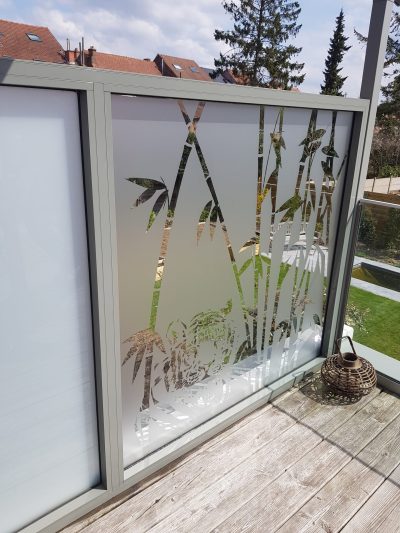 Pare-vent vitré à cadre gris d'une terrasse en bois, dont un panneau porte un film à effet sablé ajouré de tiges de bambou et d'un bouddha
