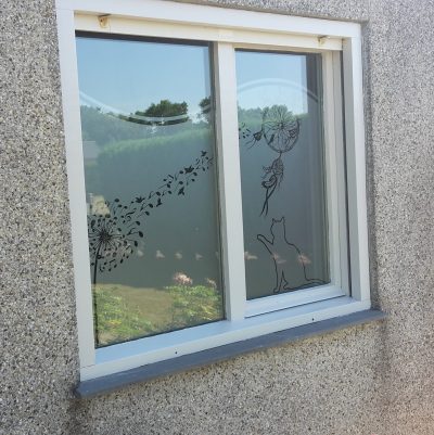 Fenêtre à deux vantaux d'une façade en crépi gravillonné avec un film à effet sablé ajouré d'un pissenlit qui s'envole en notes de musique, d'un attrape-rêves et d'un chat assis