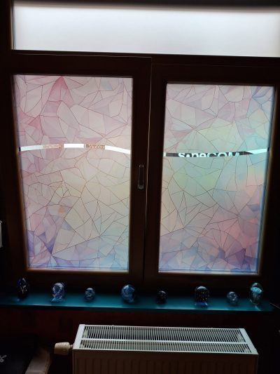Fenêtre à deux vantaux à châssis bois foncé, vue de l'intérieur, dont les deux vitrages portent un film décoratif à motif de verre éclaté en tons pastel, traversé d'une bande claire à hauteur des yeux