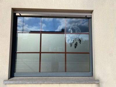 Fenêtre à croisillons d'une façade enduite beige dont le vitrage porte un film à effet sablé sur les trois rangées basses et un attrape-rêves à plumes dans l'angle supérieur droit