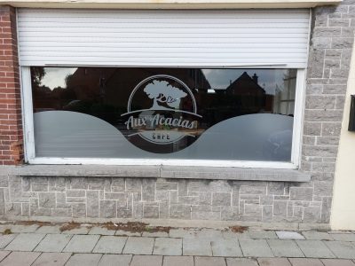 Vitrine d'un café encadrée de pierre grise, volet roulant à demi baissé, dont le vitrage porte un film à effet sablé en bandeau ondulé surmonté d'un logo à l'arbre dans un cercle
